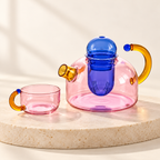 Mignone Heat-Resistant Borosilicate Glass Teapot in pink clear glass with blue infuser and amber handle, beside matching cup