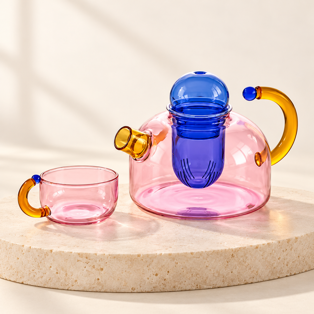 Mignone Heat-Resistant Borosilicate Glass Teapot in pink clear glass with blue infuser and amber handle, beside matching cup